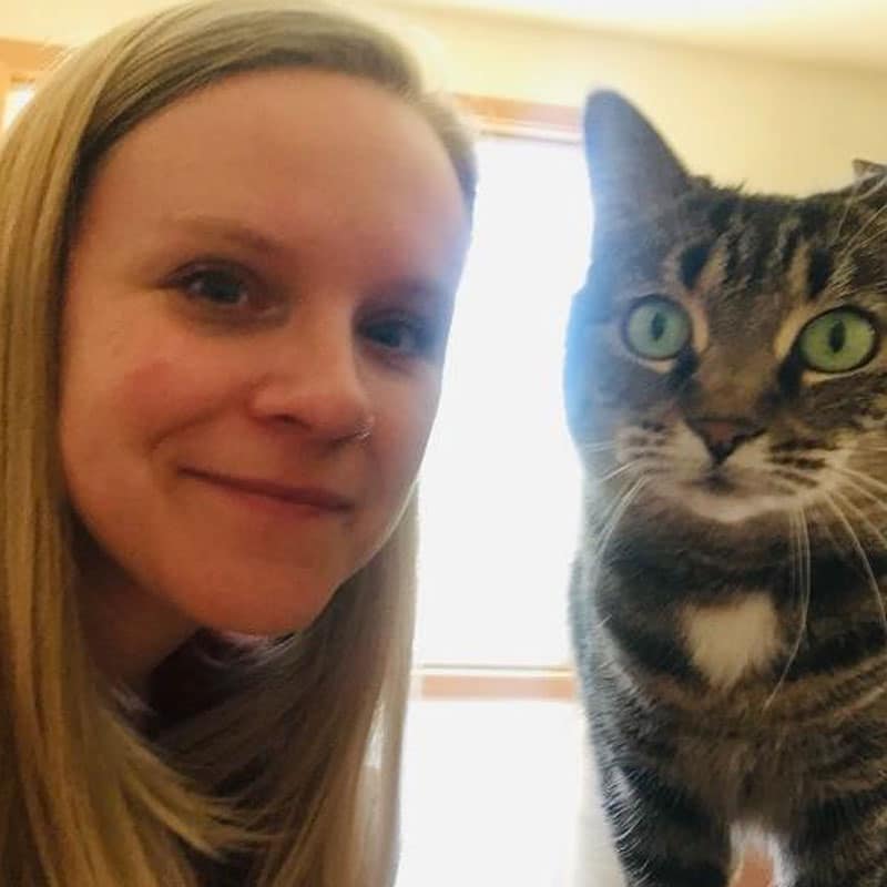 A woman with long blonde hair smiles at the camera beside a tabby cat with green eyes. They are indoors, with a sunlit window in the background.